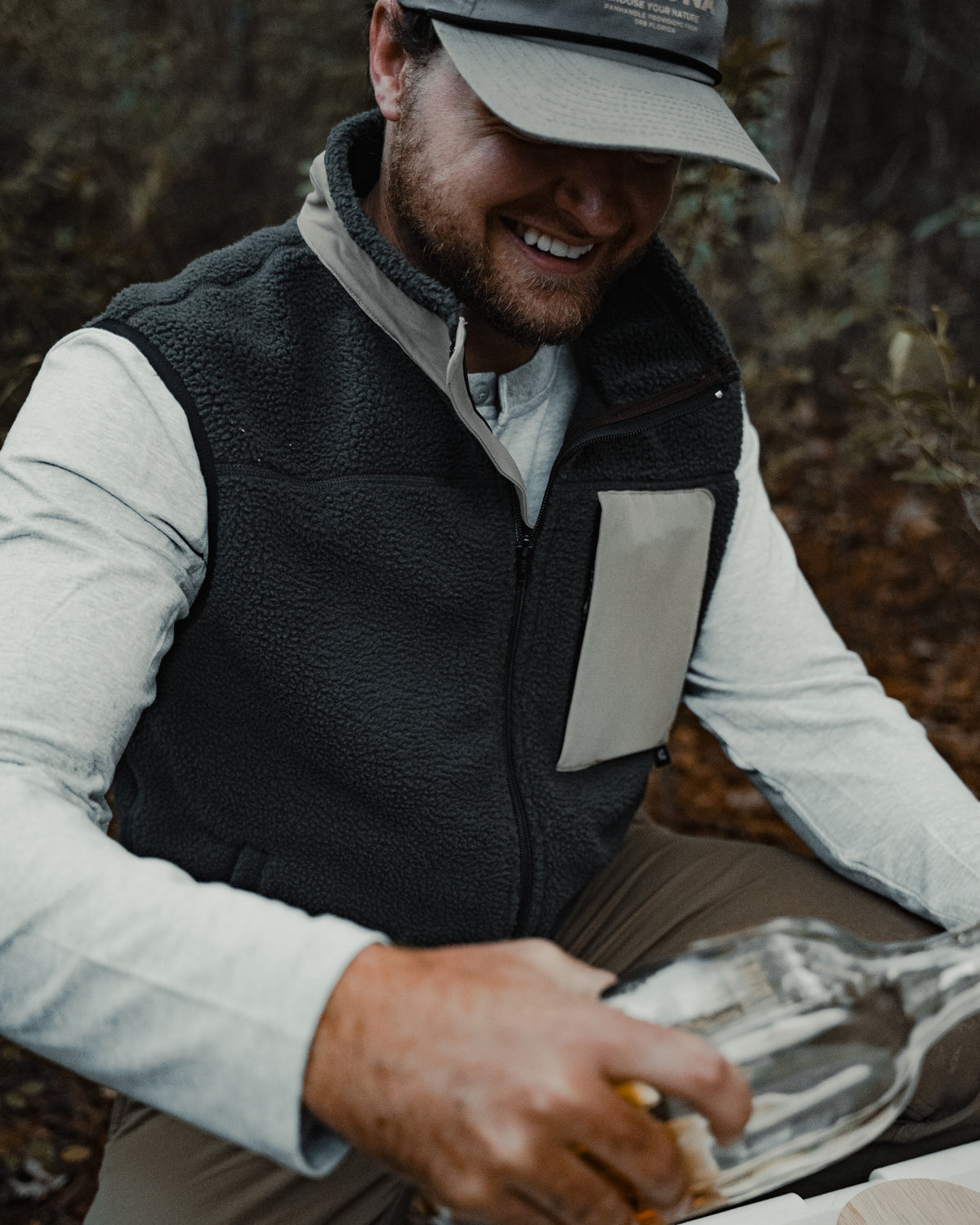 A man in a gray cap and Choona Pt. Washington Sherpa Vest smiles while pouring a drink outdoors amid greenery. The high-pile fleece of the vest delivers cozy warmth for all your adventures.