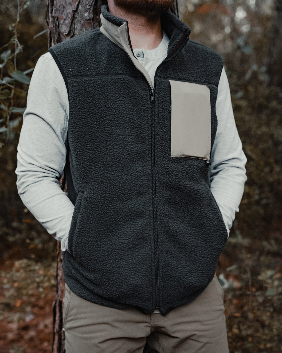 A person in a Choona Pt. Washington Sherpa Vest with a contrasting chest pocket, light long-sleeve shirt, and khaki pants leans against a tree outdoors, hands in pockets.