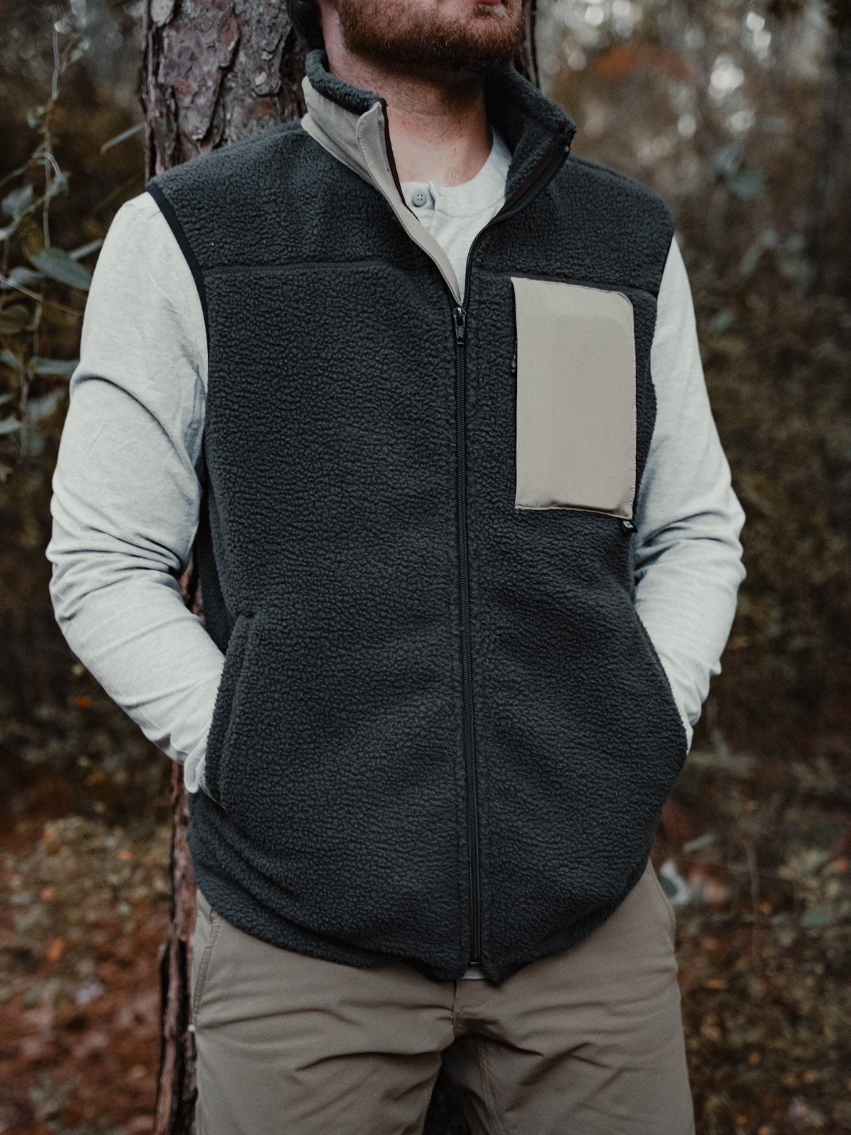 A person in a Choona Pt. Washington Sherpa Vest with a contrasting chest pocket, light long-sleeve shirt, and khaki pants leans against a tree outdoors, hands in pockets.