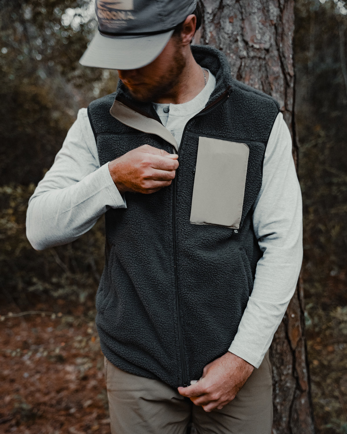 A man in a Choona Pt. Washington Sherpa Vest with Durable Water Repellent finish, a light long-sleeve shirt, and a cap stands by a tree, zipping his vests chest pocket as he looks down, his face partially hidden.