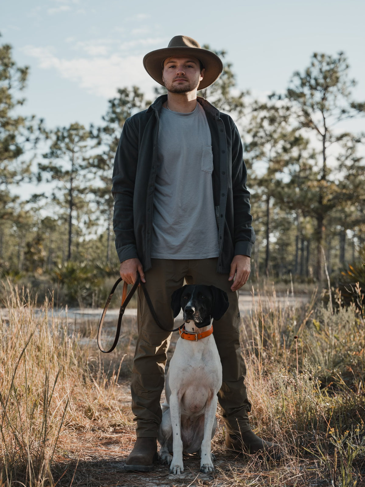 A man in outdoor clothes and a Choona Skipper Shacket stands in a grassy field holding a leash, with his black and white dog sitting in front. Pine trees and blue sky suggest crisp, cold weather in the background.
