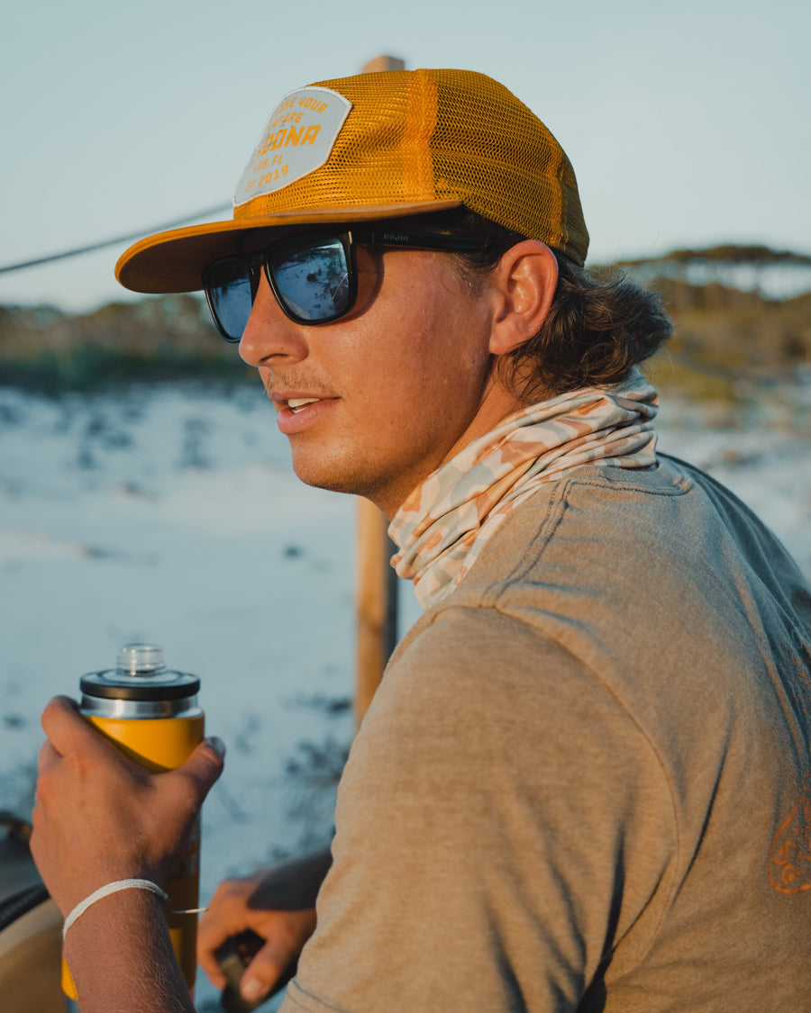 A young man in a yellow cap, sunglasses, and Choonas Grayton Sun Mask holds a yellow insulated bottle outdoors near the beach, with blurred sand and greenery in the background.