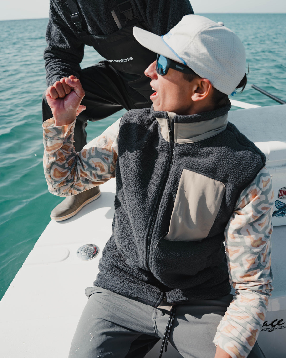 A person wearing the Choona Pt. Washington Sherpa Vest, sunglasses, and a white cap sits on a boat, gesturing while talking. Another person in dark clothing stands nearby as the boat floats on calm blue water under a clear sky.
