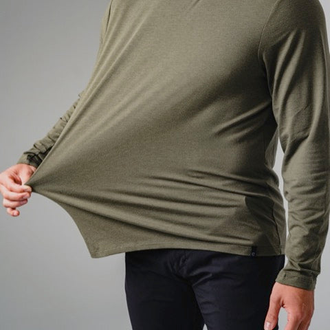 A person wearing a long-sleeve olive green shirt and black pants stretches the side of their shirt outward, demonstrating the fabrics stretchiness. The persons head is not visible in the image.