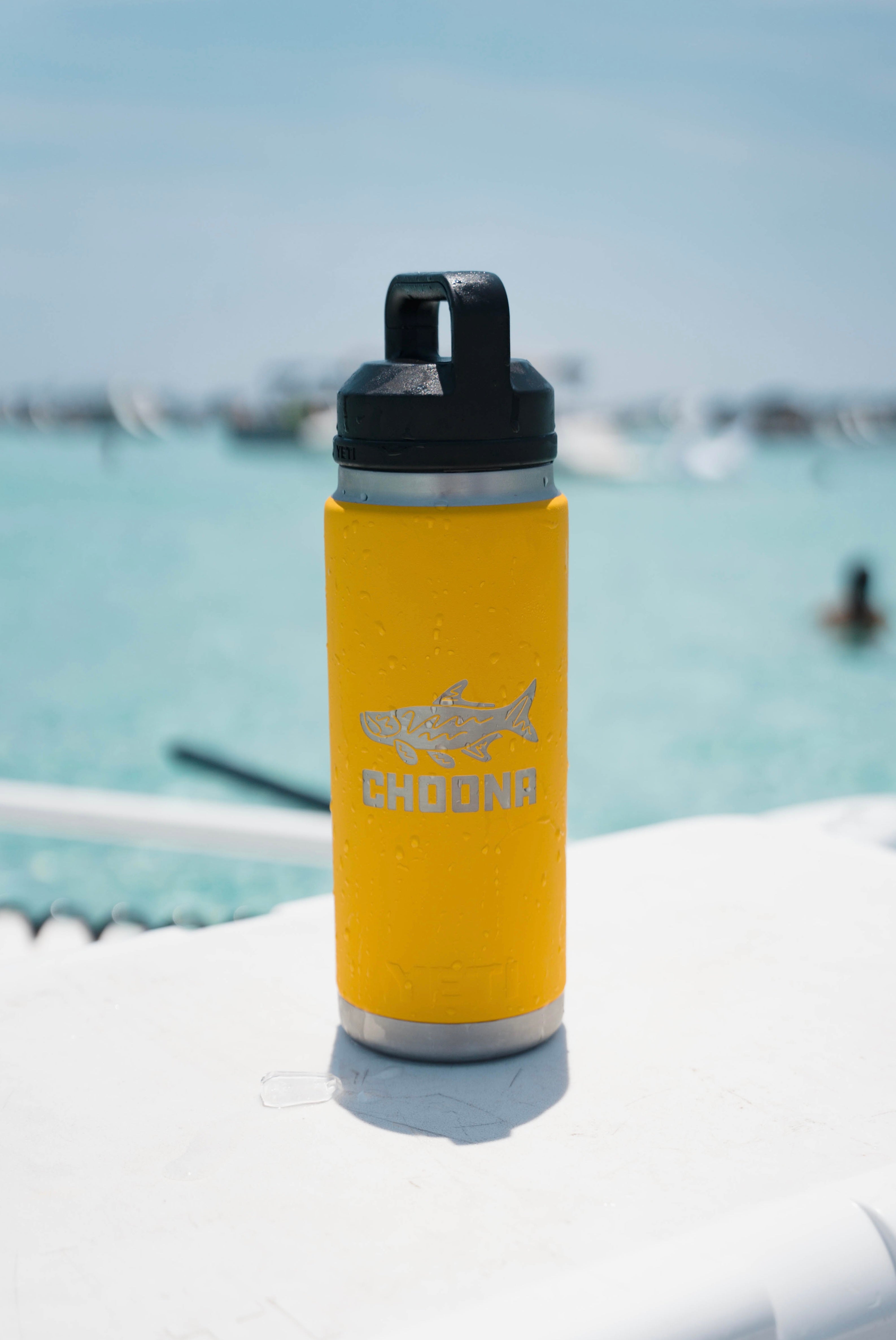 A yellow Choona YETI® X CHOONA RAMBLER® 26oz insulated bottle with CHOONA and a fish graphic, featuring a chug spout lid, rests on a white surface near blue water, with people and boats blurred in the background.