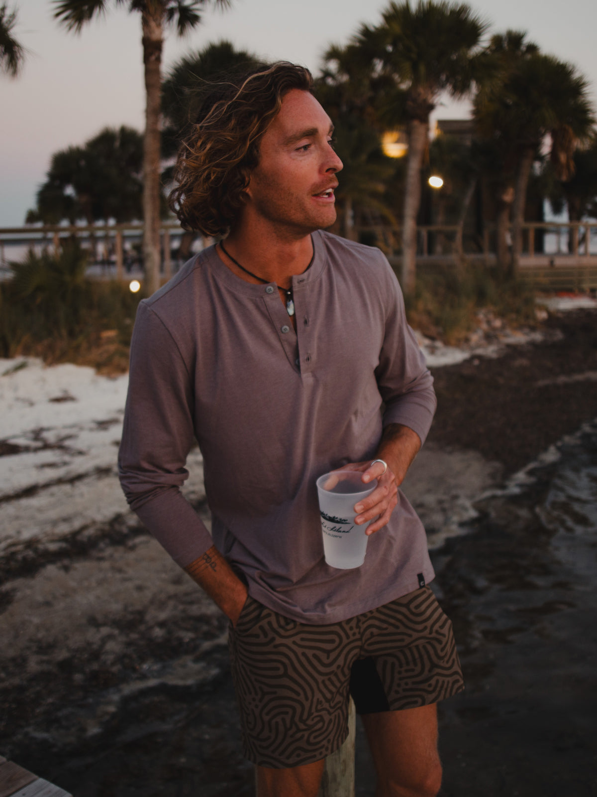 A man with wavy hair stands outdoors by water and palm trees at sunset, holding a translucent cup and wearing Choonas Versatile LS Henley paired with patterned shorts.