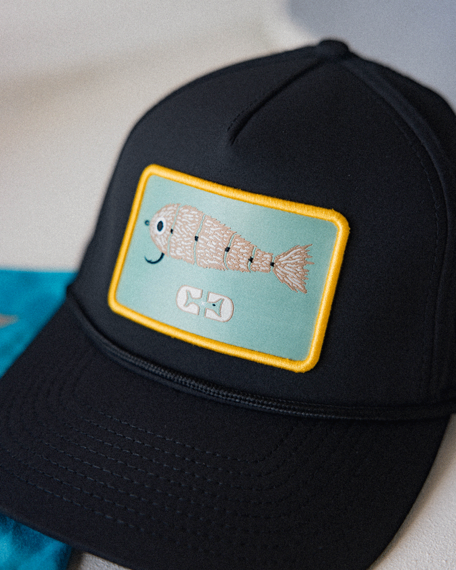 A close-up of the Choona X Dent Redfish Snapback by Choona, a black cotton twill cap with a rectangular patch showing an embroidered smiling fish and two fishhooks, all outlined in yellow.