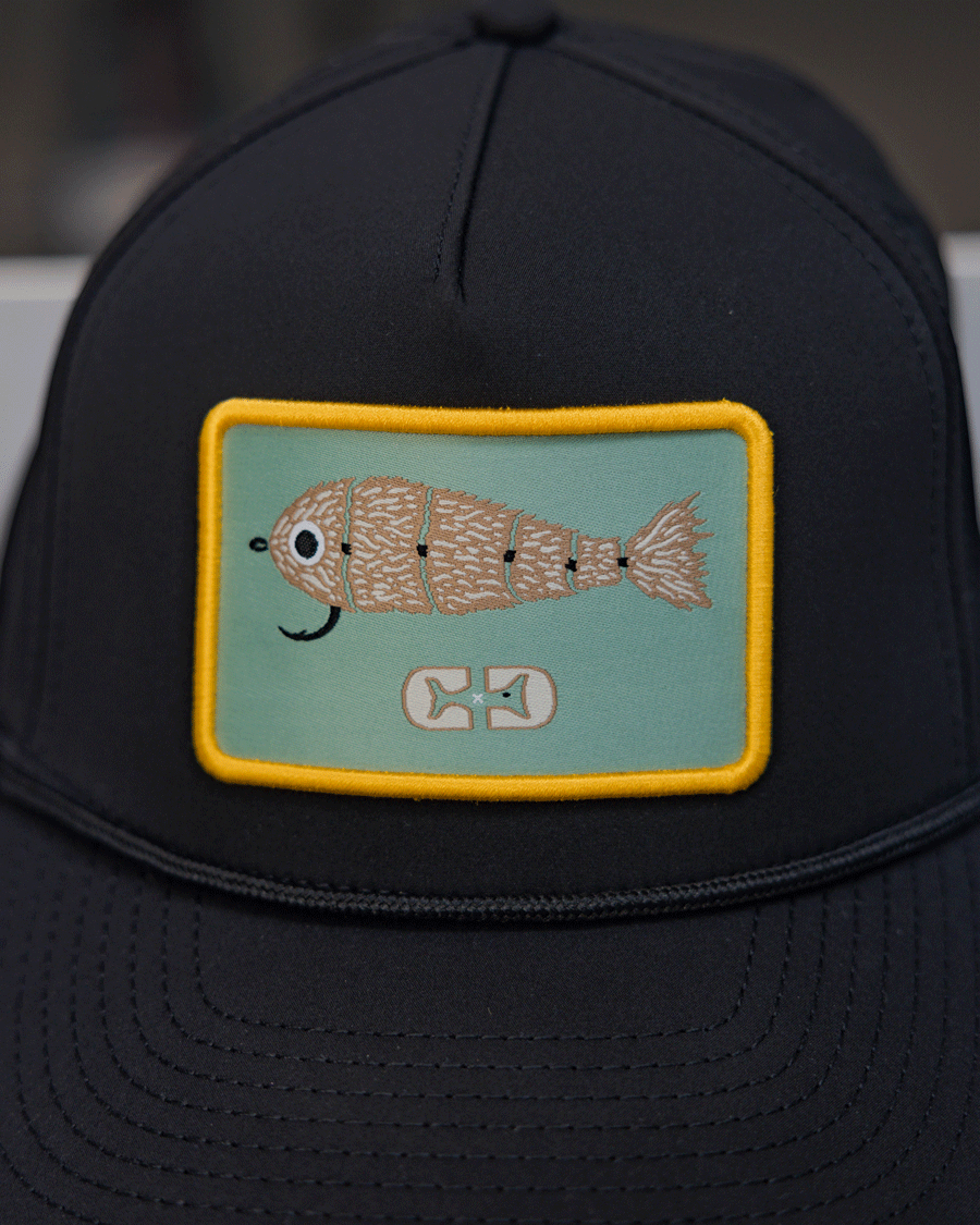 The Choona X Dent Redfish Snapback by Choona is a black cotton twill cap with a rectangular patch showing a cartoon fish with scales and a hook-shaped mouth on light blue, bordered in yellow.
