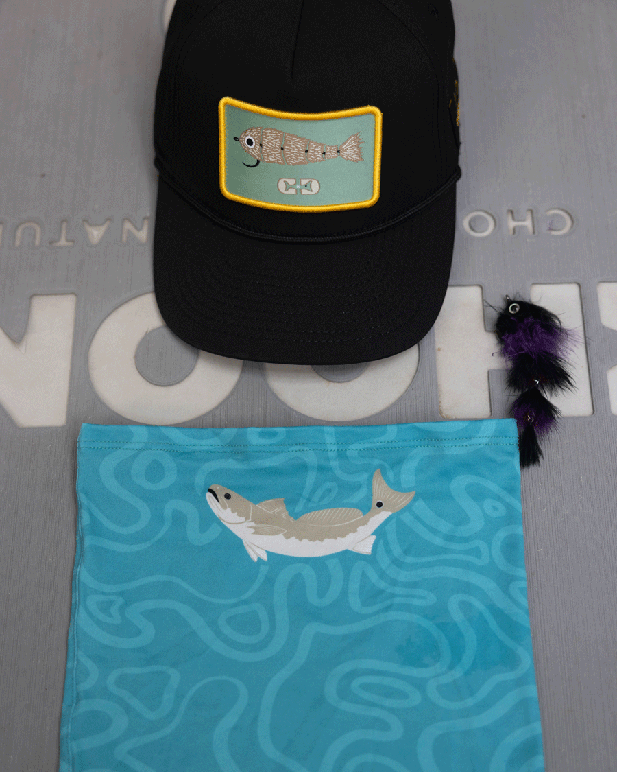 A black cap with a rectangular fish patch, a blue Choona Grayton Sun Mask made from ultra-breathable fabric with a fish graphic, and a purple and black fishing lure are shown on a gray surface with partially visible white text.