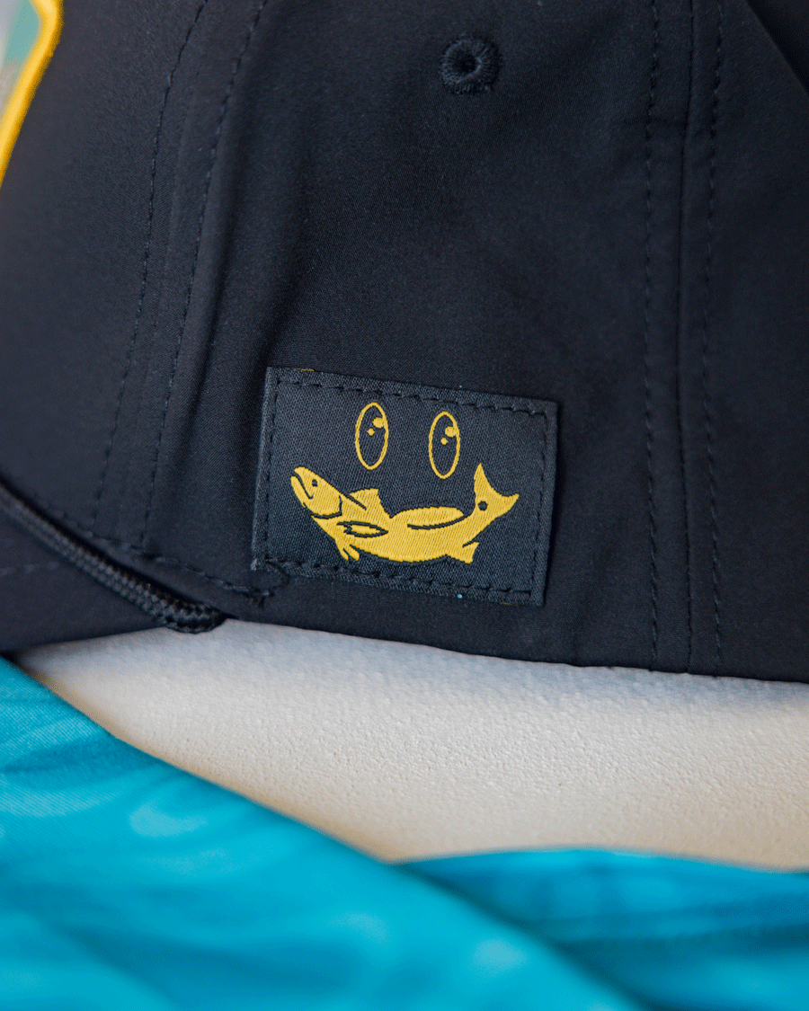 A close-up of the Choona X Dent Redfish Snapback by Choona, a black cotton twill hat with a small stitched patch showing a yellow smiling fish and two circles above it. Blue fabric appears in the foreground.