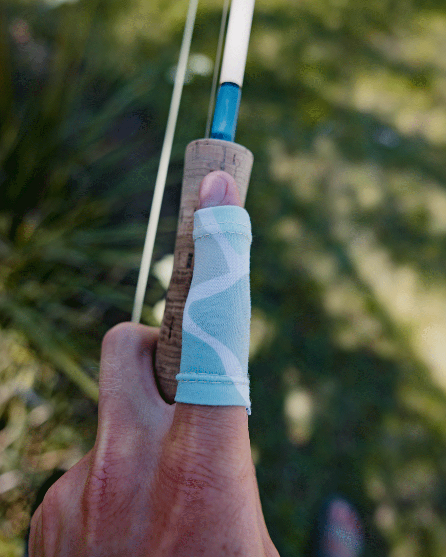 A hand wrapped with a Choona Strip Guard protects against line burn while holding a fishing rod. The grassy, blurred background hints at an ideal outdoor setting for fly anglers.