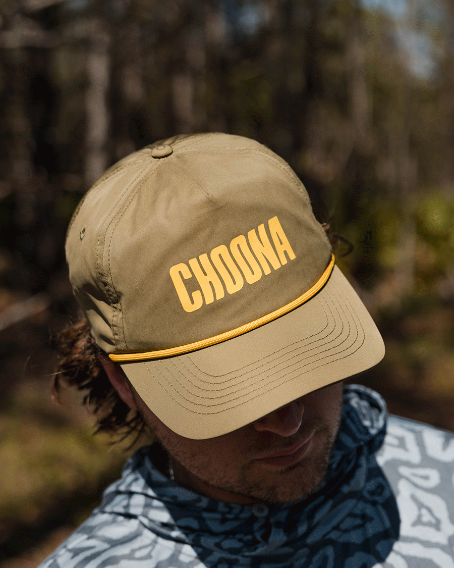 On a coastal adventure, someone sports the Choona Wavy Snapback—a khaki quick-drying cap with bold yellow CHOONA lettering—paired with a patterned gray hoodie as the brim shades their face.