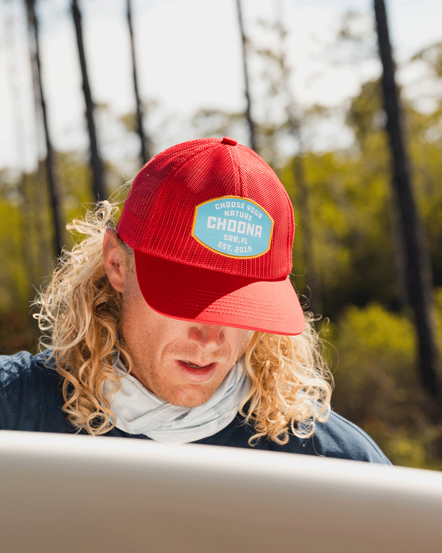 Someone with long, curly blonde hair wears a Choona Breezy Snapback - Red and a light neck gaiter while looking down at a surfboard outdoors, with trees and sunlight in the background.