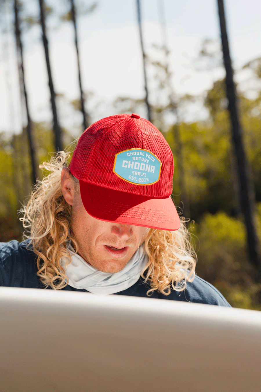 Someone with long, curly blonde hair wears a Choona Breezy Snapback - Red and a light neck gaiter while looking down at a surfboard outdoors, with trees and sunlight in the background.