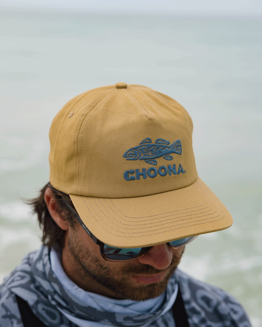 A man wearing sunglasses, a gray patterned neck gaiter, and a mustard yellow Choona Redfish Snapback stands by the water, looking down.