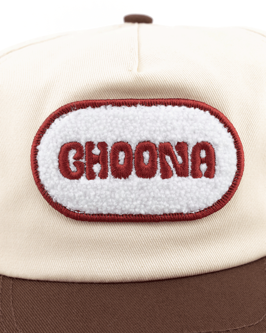 The Choona Fly Patch Snapback is a cream cotton twill cap with a brown brim, featuring a white oval patch outlined in red and embroidered with CHOONA in bold red letters.