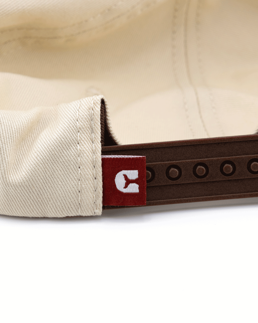 Close-up of the Choona Fly Patch Snapback, an unstructured beige cotton twill cap with a brown adjustable snapback and a red tag featuring a white stylized C logo sewn near the back closure.