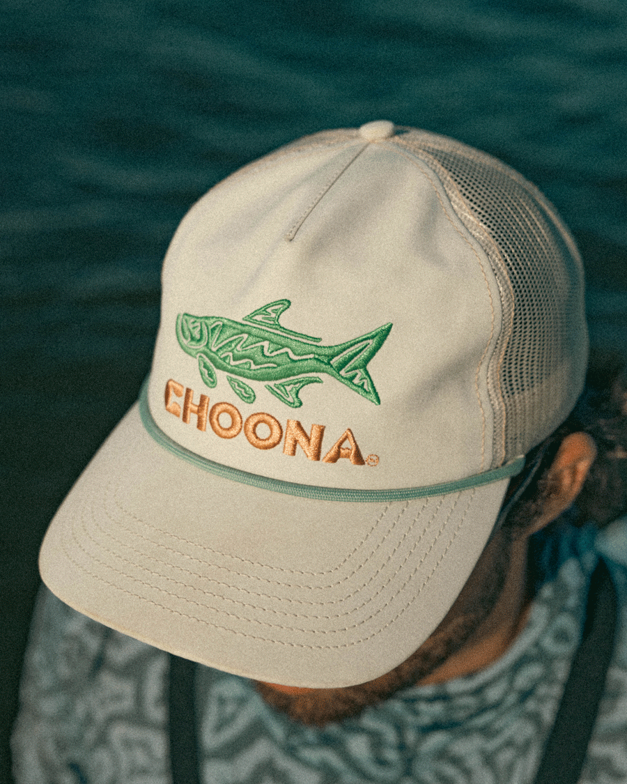 Someone near the water wears a beige Choona Silver King Snapback hat with a mesh back, featuring a green and blue fish plus the Choona tarpon logo embroidered on the front.