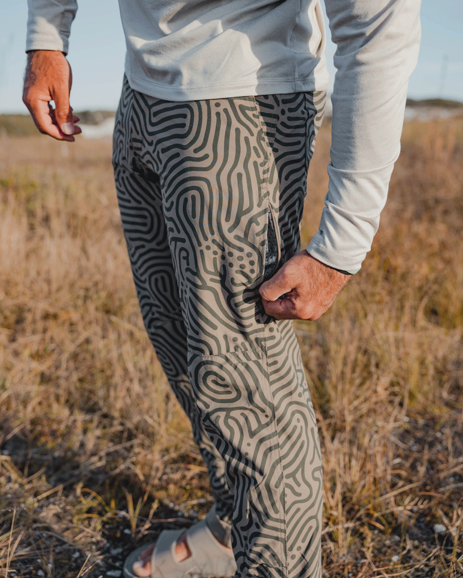 Wearing Choona Drifter Pant with a map-like pattern, four-way stretch comfort, a light long-sleeve shirt, and sandals, a person pulls the side pocket zipper outdoors against dry grass and a blurred landscape.