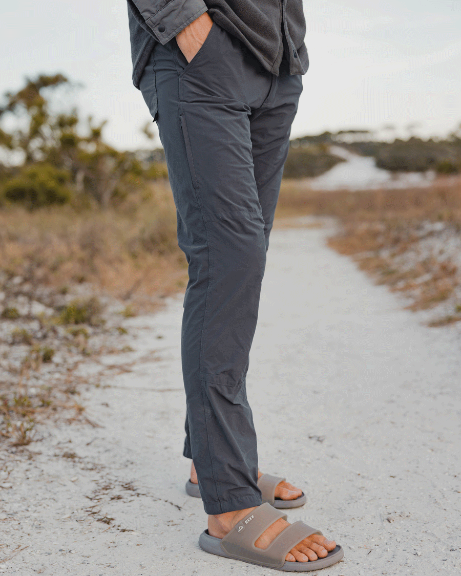 A person stands outdoors on a sandy path, wearing the Choona Drifter Pant with four-way stretch, a matching jacket, and tan slide sandals. Their hands are in their pockets, surrounded by dry grass and trees.