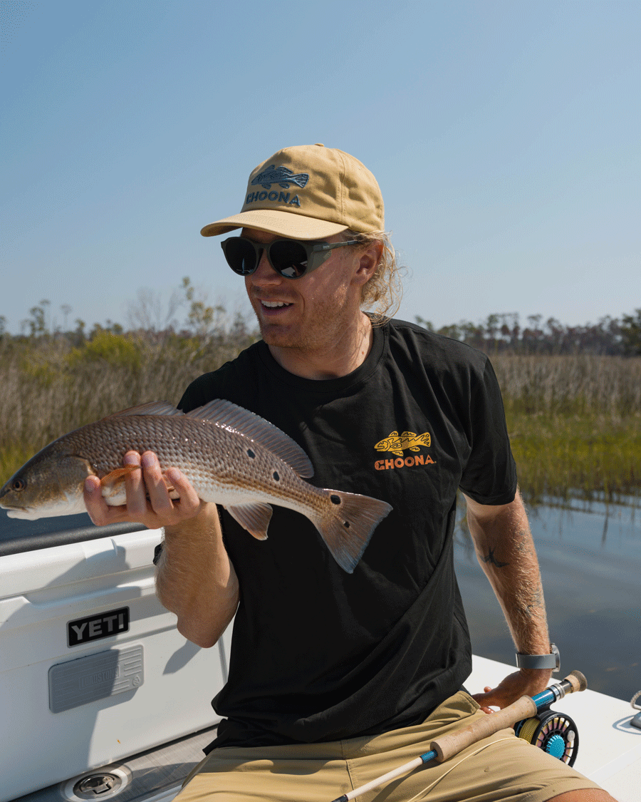 A man in sunglasses and a tan cap sits on a boat, smiling as he holds a fish. He wears the Choona Redfish Graphic Tee with tan shorts. Marshland and trees extend behind him under the clear sky.