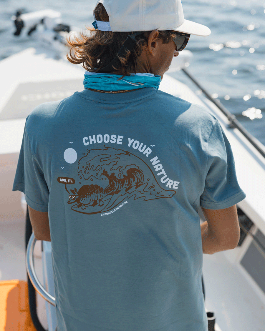 A person stands on a boat wearing the Choona Armachillo Graphic Tee in blue, featuring "CHOOSE YOUR NATURE" and an illustrated wave, gazing at the water on a sunny day.