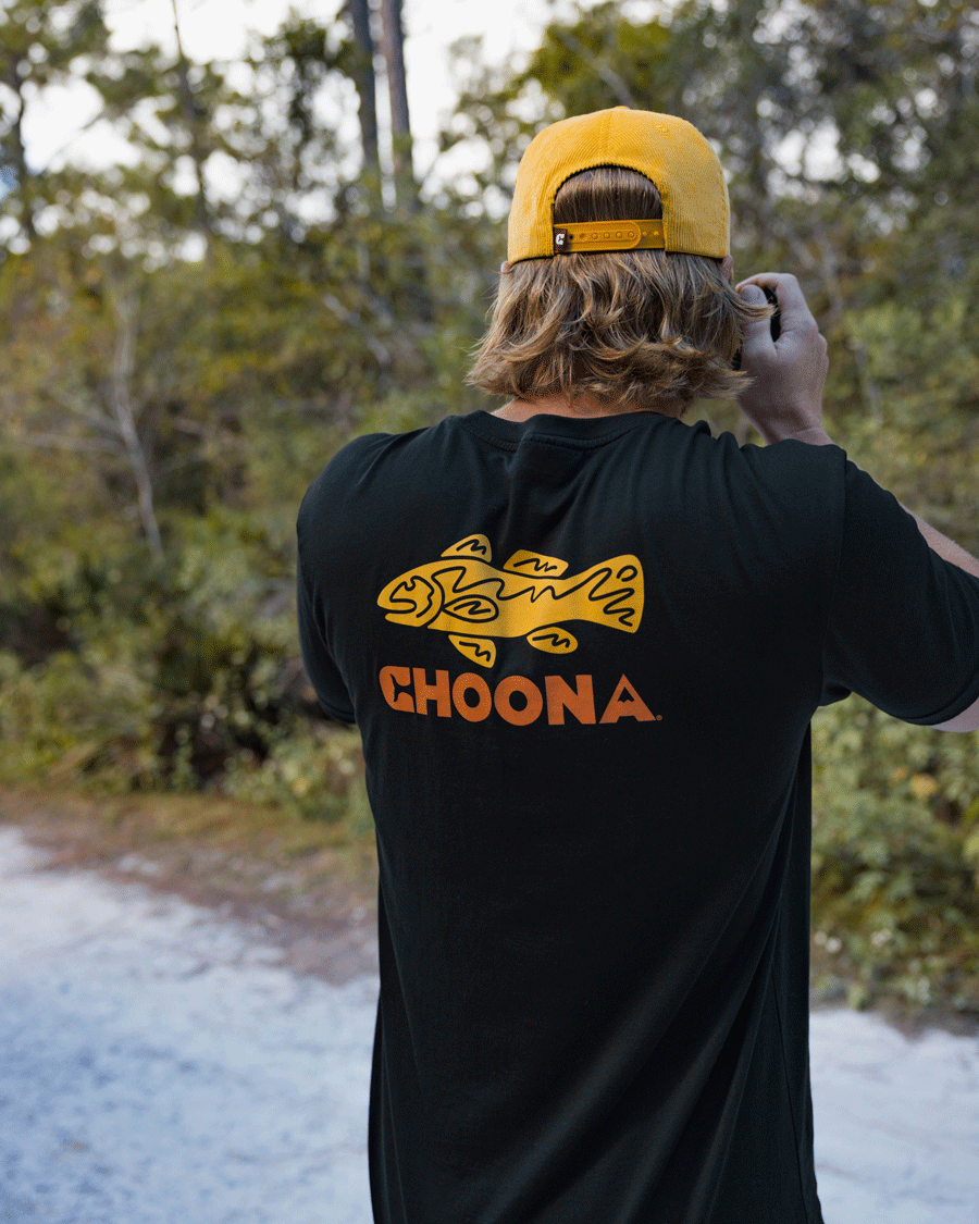 A person with long blond hair wears a yellow cap backwards and a Choona Redfish Graphic Tee, standing outdoors taking a photo in nature.