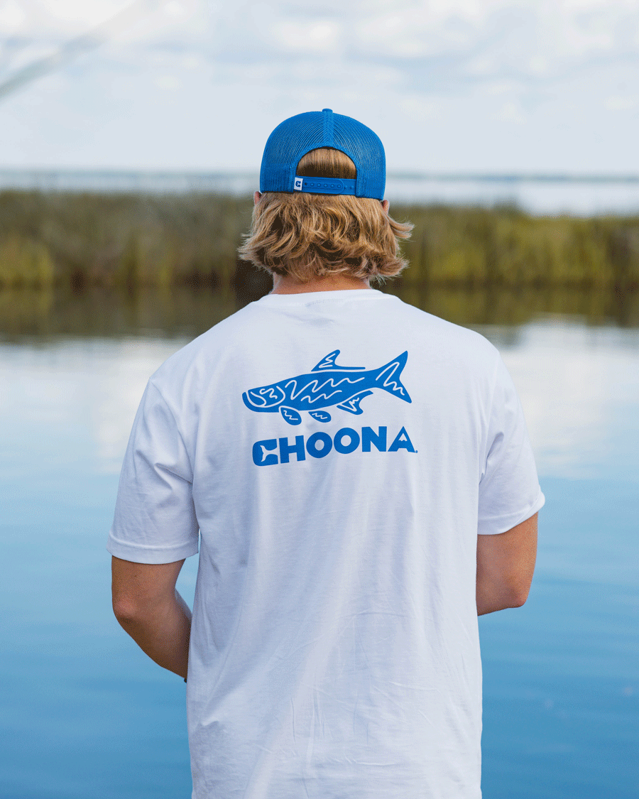 A person with blond hair, wearing a blue cap and a white Choona Tarpon Graphic Tee, stands by calm water and marsh grasses, facing away from the camera.