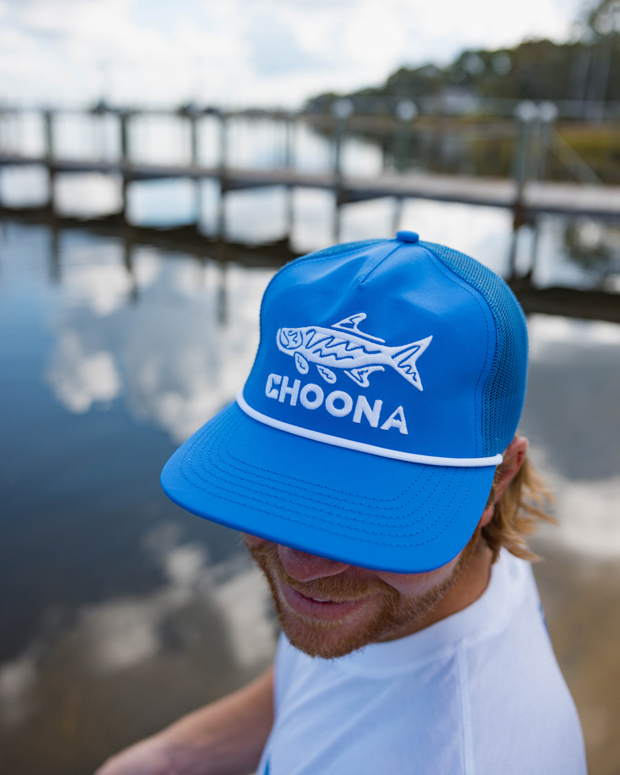 A person wearing a blue Choona Silver King Snapback hat with a cartoon fish and "CHOONA" stands by a calm waterfront near a wooden dock. This breathable mesh snapback is perfect for relaxed days by the water.