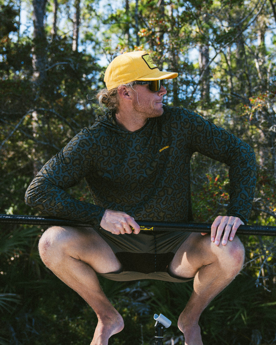A man in a Choona Grayton Ultra-Lightweight Hoodie, yellow cap, and sunglasses squats outdoors holding a paddle, surrounded by trees and foliage.