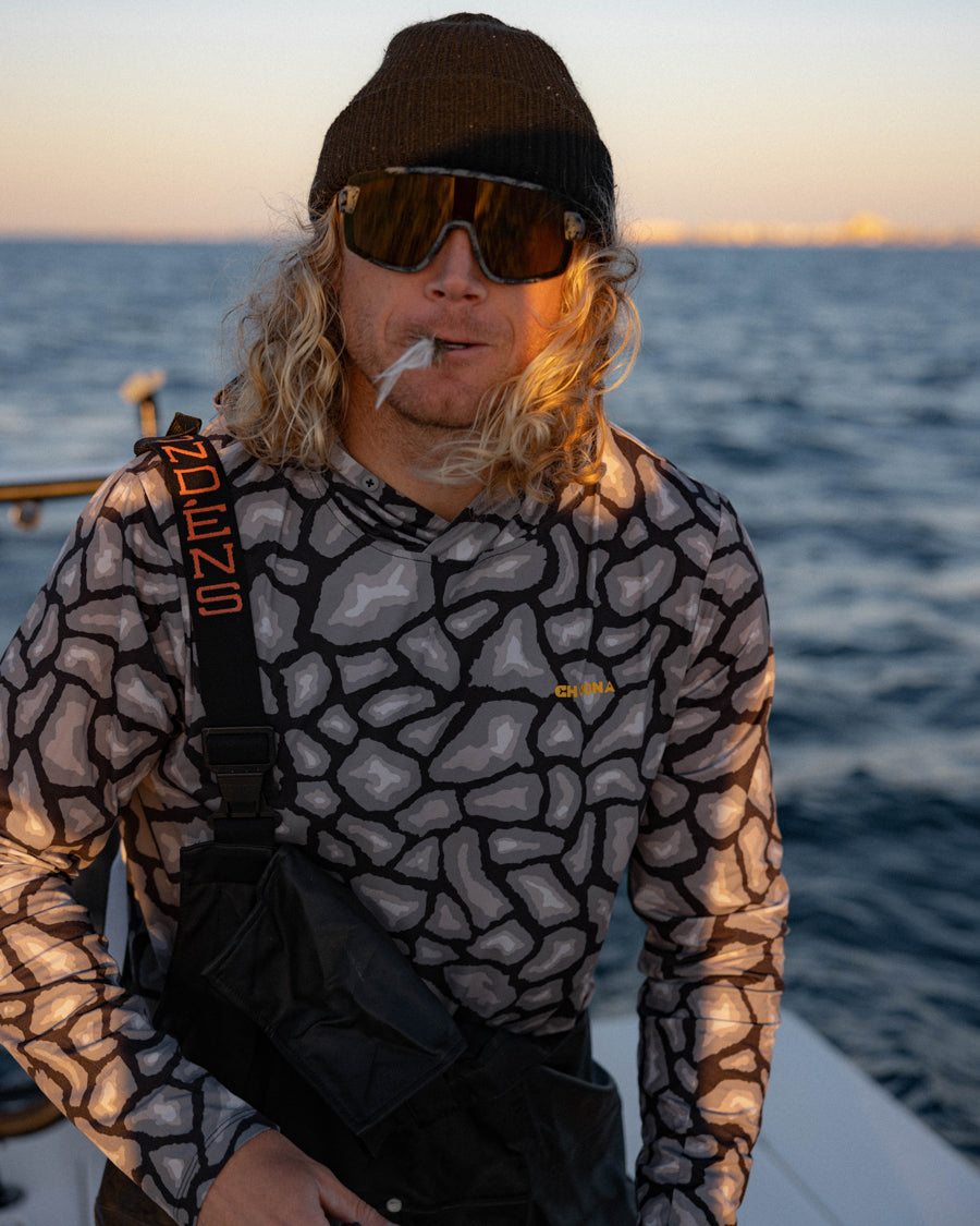 A person with long blond hair, in a black beanie, large sunglasses, and a patterned shirt stands on a boat with water behind them, holding a cigarette and layering with the Choona Grayton Ultra-Lightweight Hoodie for moisture management.