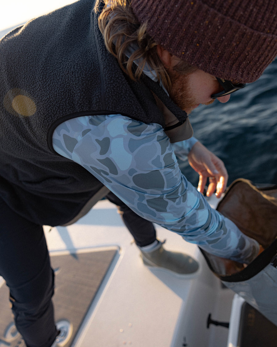A person in a Choona Grayton Ultra-Lightweight Hoodie with camo sleeves, a knit beanie, and sunglasses stands on a boat, reaching into a bag as sunlight highlights the scene.