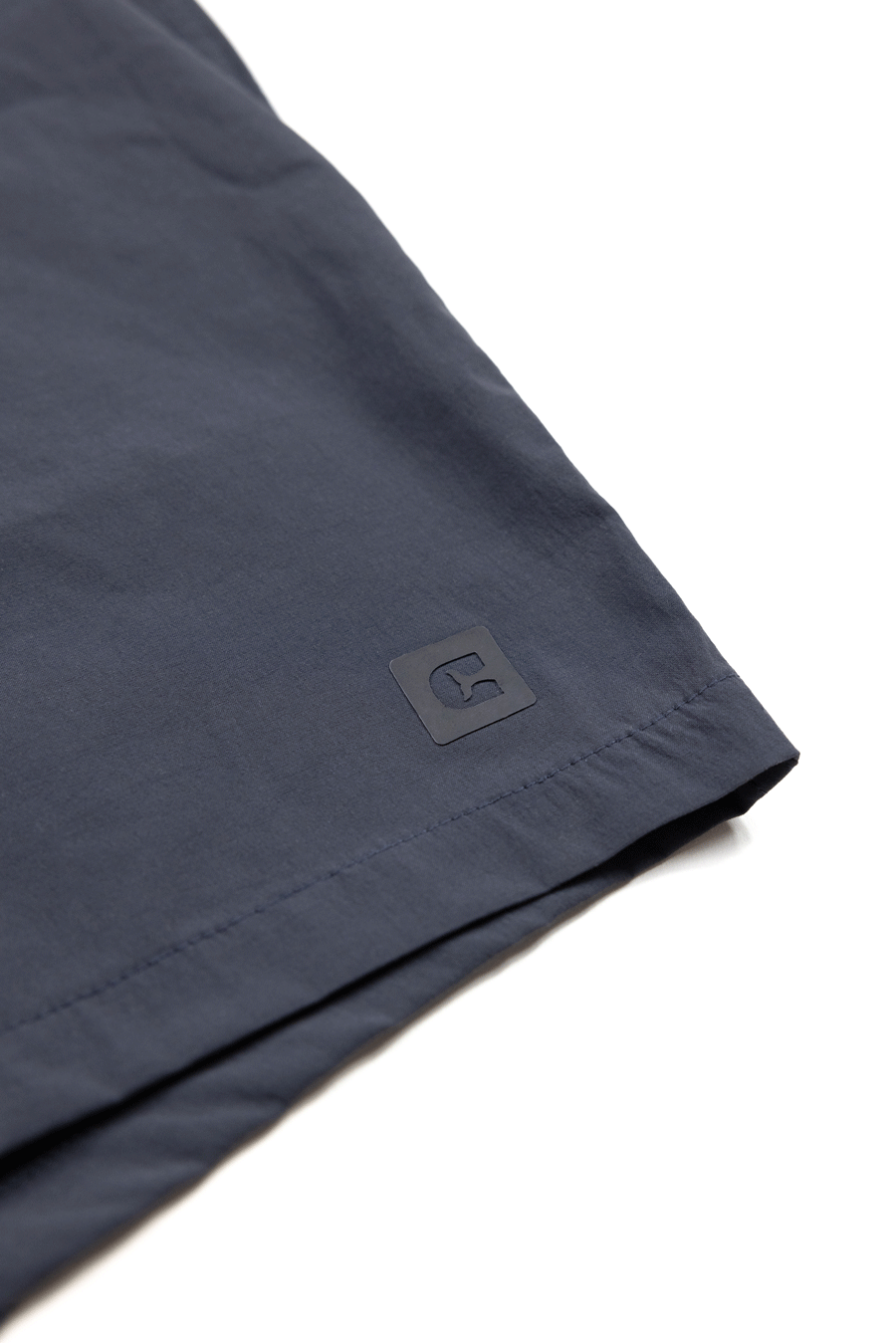 Close-up of the hem of Choonas Shore Short in dark blue, showcasing a small square black rubber logo patch—perfect for coastal adventures with advanced moisture-wicking technology.