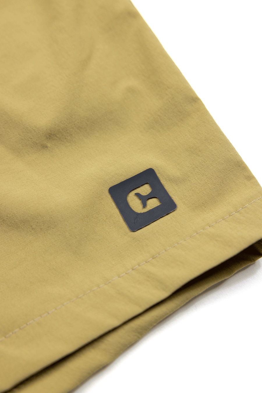 Close-up of the light tan Choona Shore Short hem, showcasing a small black square logo with a stylized “C”—ideal for coastal activities due to advanced wicking technology.