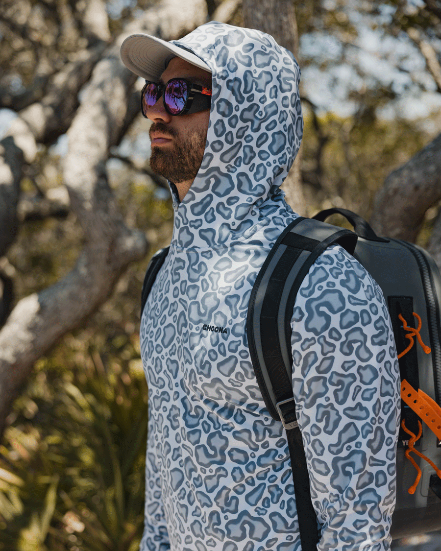 A man stands outdoors among trees, looking ahead, wearing the Choona Grayton Ultra-Lightweight Hoodie for breathable performance, sunglasses, a cap, and a backpack.