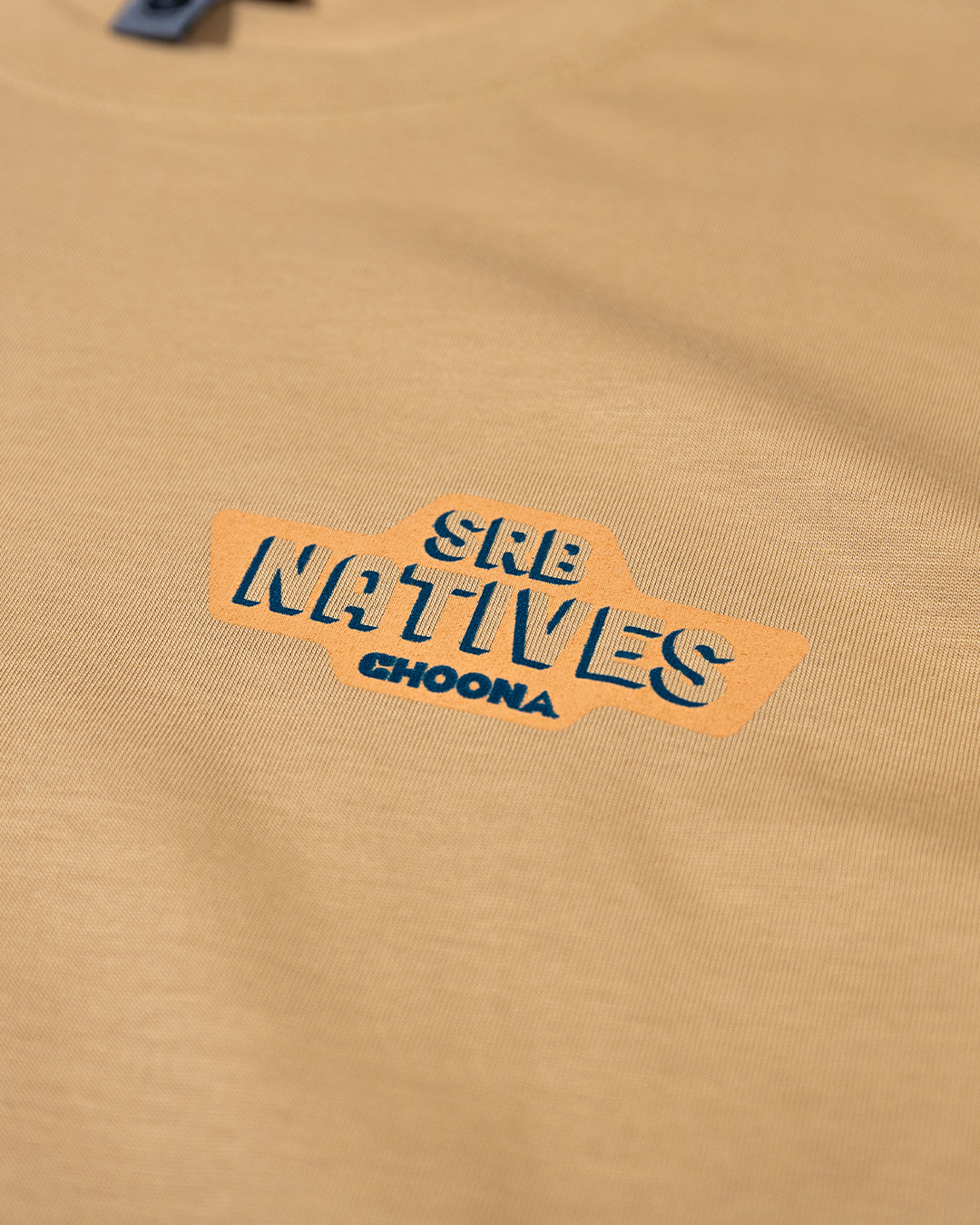 A close-up of the Choona SRB Natives Graphic Tee, a tan Pima Cotton shirt featuring SRB NATIVES CHOONA in bold blue letters atop an orange background—ideal for Santa Rosa Beach style.