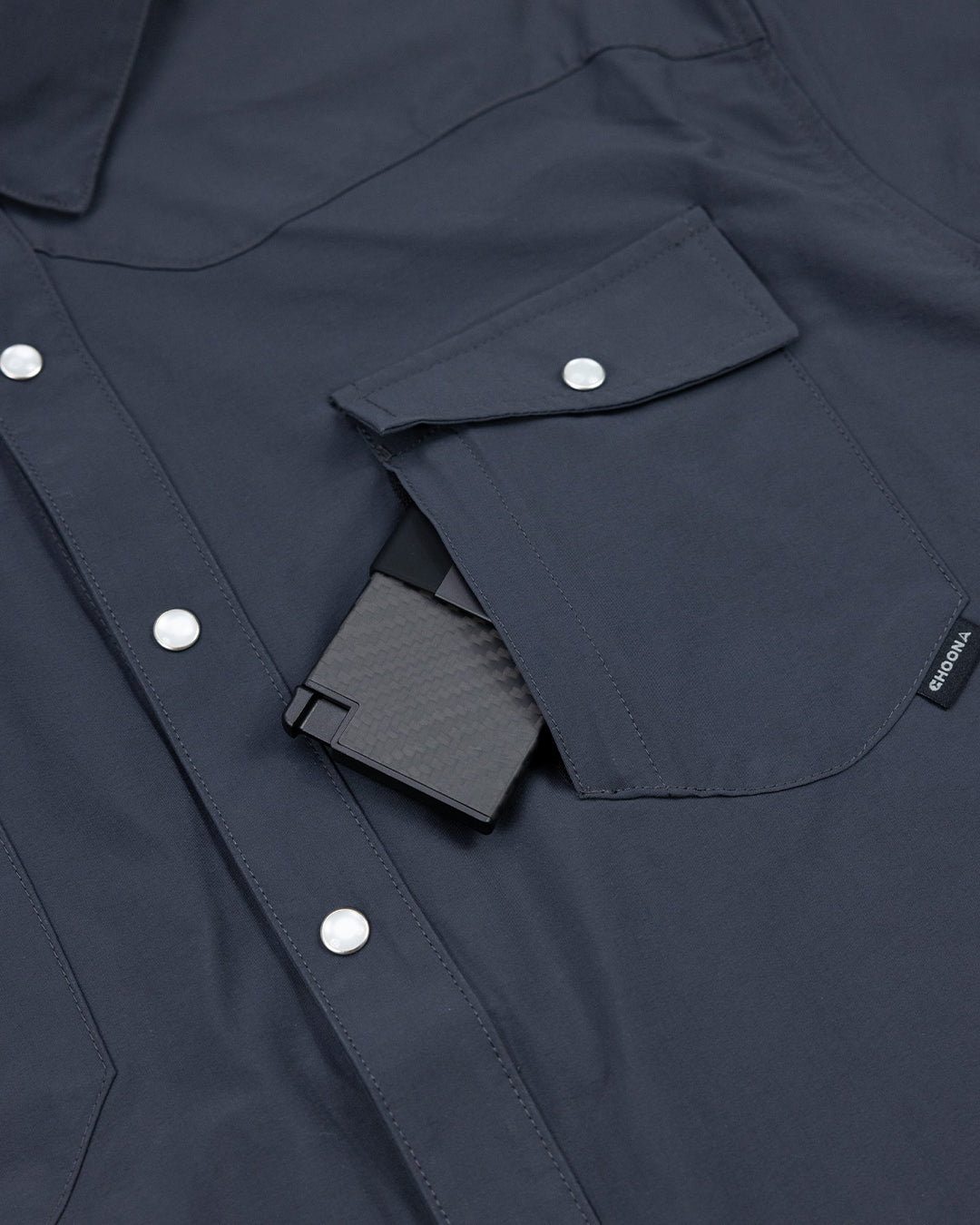 A close-up of a dark blue Choona Panhandle Pearl Snap shirt with white buttons and a front pocket, partially showing a black textured wallet—perfect for that coastal cowboy style.