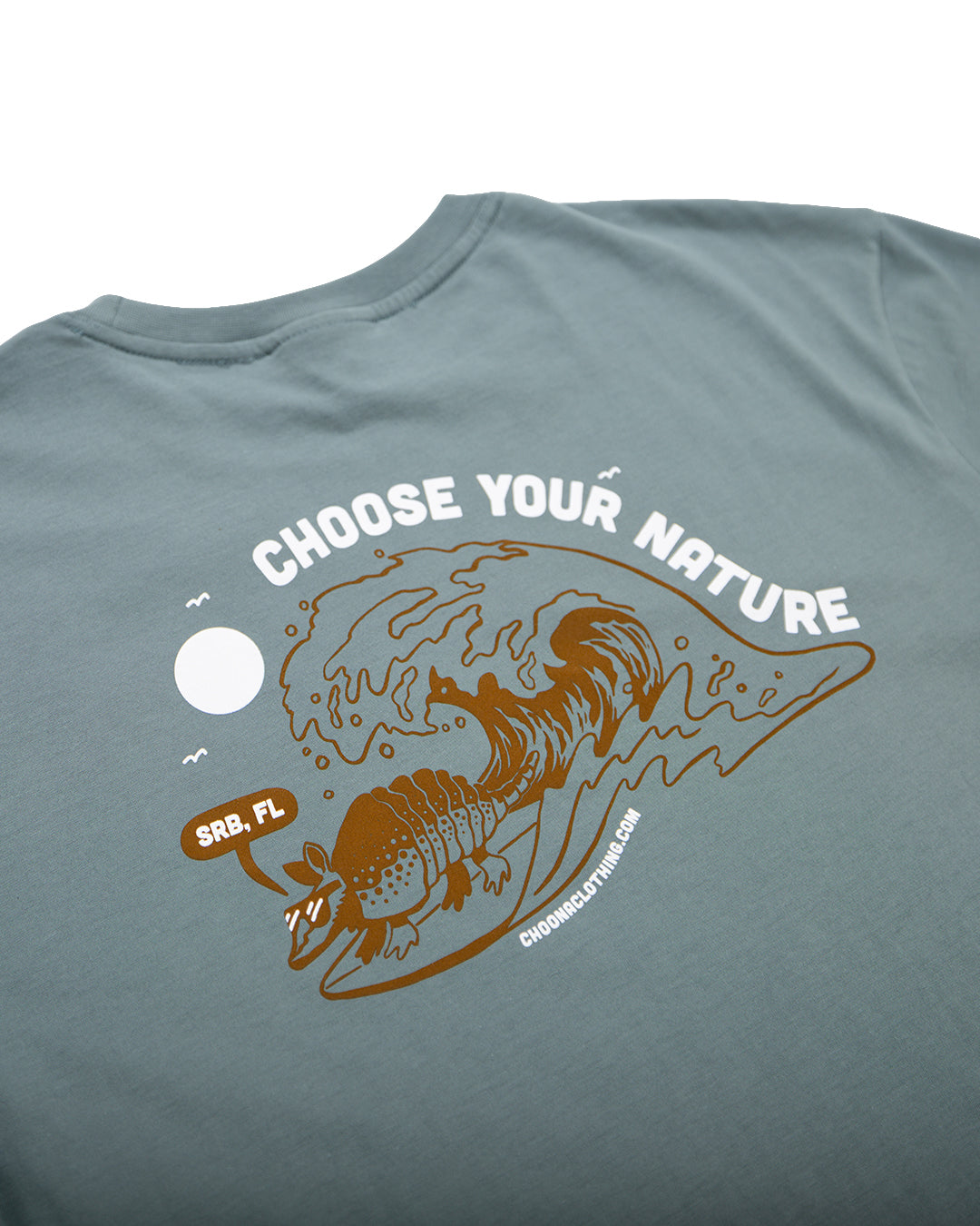 The Choona Armachillo Graphic Tee is a retro blue-green surf shirt with a surfing armadillo and "CHOOSE YOUR NATURE" over SRB, FL in a bubble. Made of USA-grown cotton, it also features CHOOSEYOURNATURE.COM under the graphic.