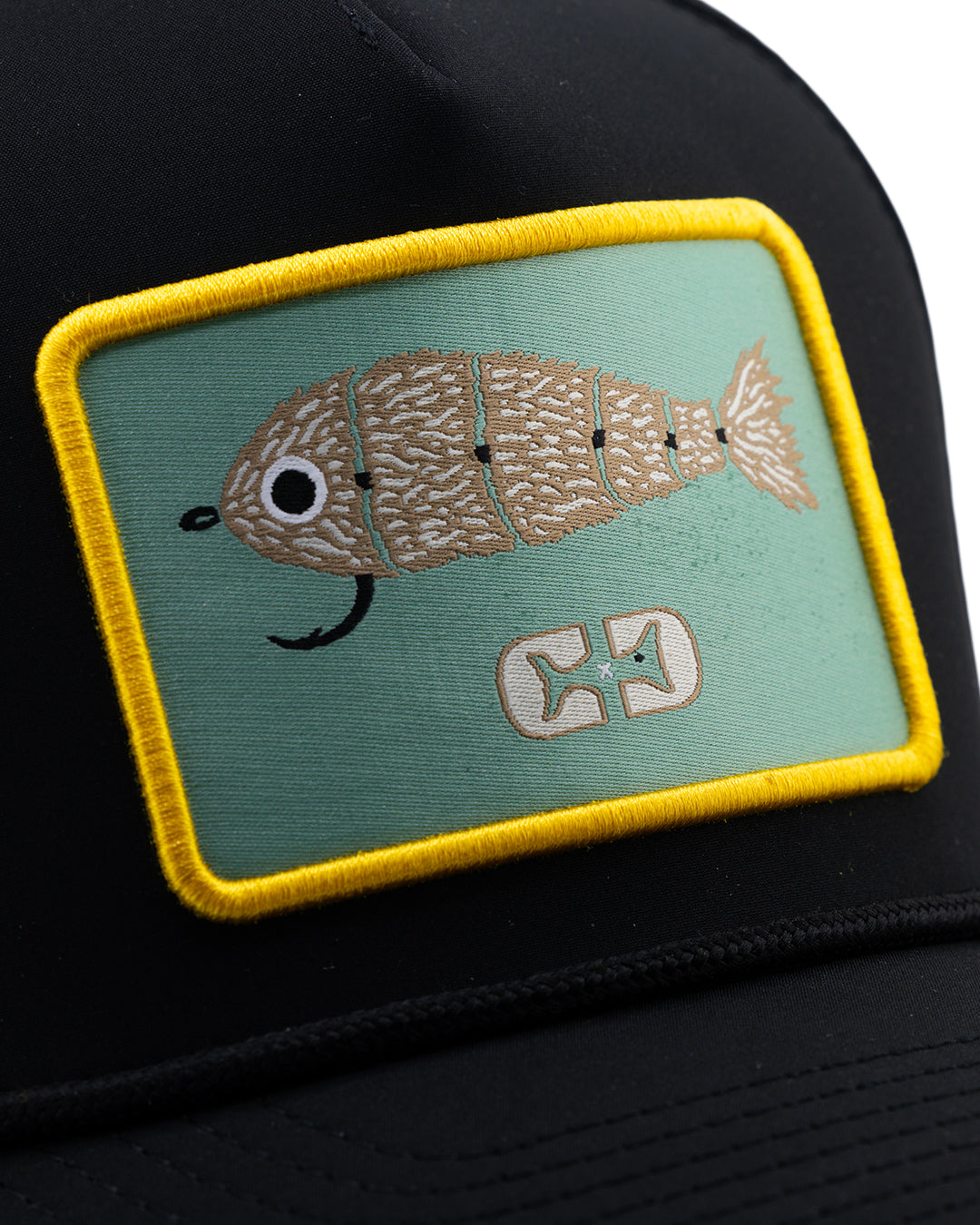 The Choona X Dent Redfish Snapback by Choona is a black hat with a rectangular yellow-edged patch featuring a tan and white cartoon fish on green, designed with fish bones and a white symbol below.