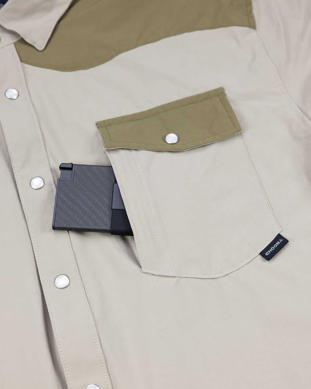 A beige Choona Panhandle Pearl Snap with an olive green pocket flap channels coastal cowboy style. A black, foldable smartphone peeks from the chest pocket, accented by a small black CHOONA tag on the edge.