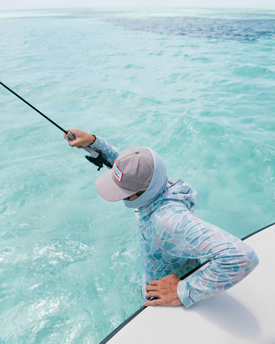 Wearing the Choona Grayton Ultra-Lightweight Hoodie and a cap, a person stands on a boat with a fishing rod above clear turquoise water. The breathable fabric keeps them cool as they look down, facing away from the camera.