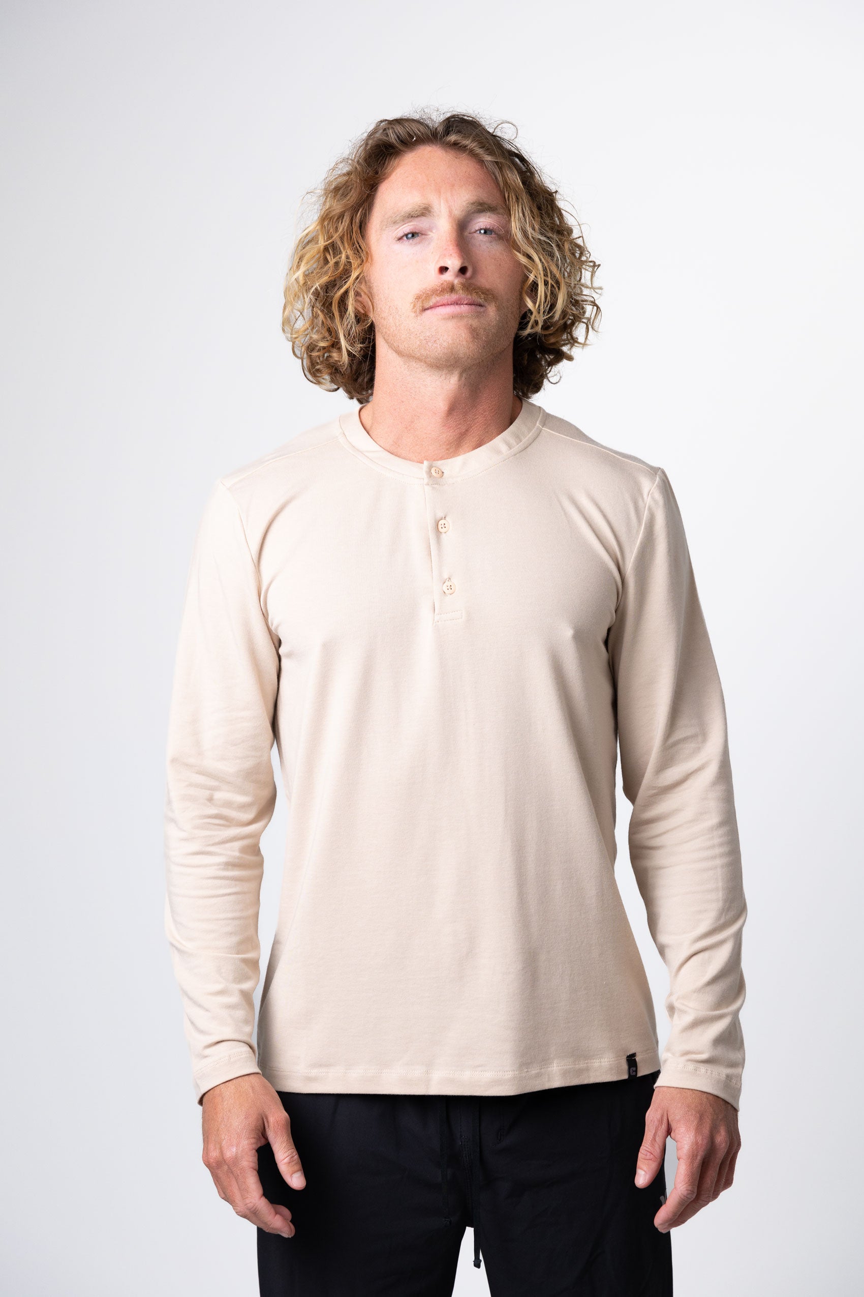 A man with curly blond hair wears Choonas Versatile LS Henley in beige and black pants, standing against a plain light background—showcasing an outleisure essential ideal for an active outdoor lifestyle.
