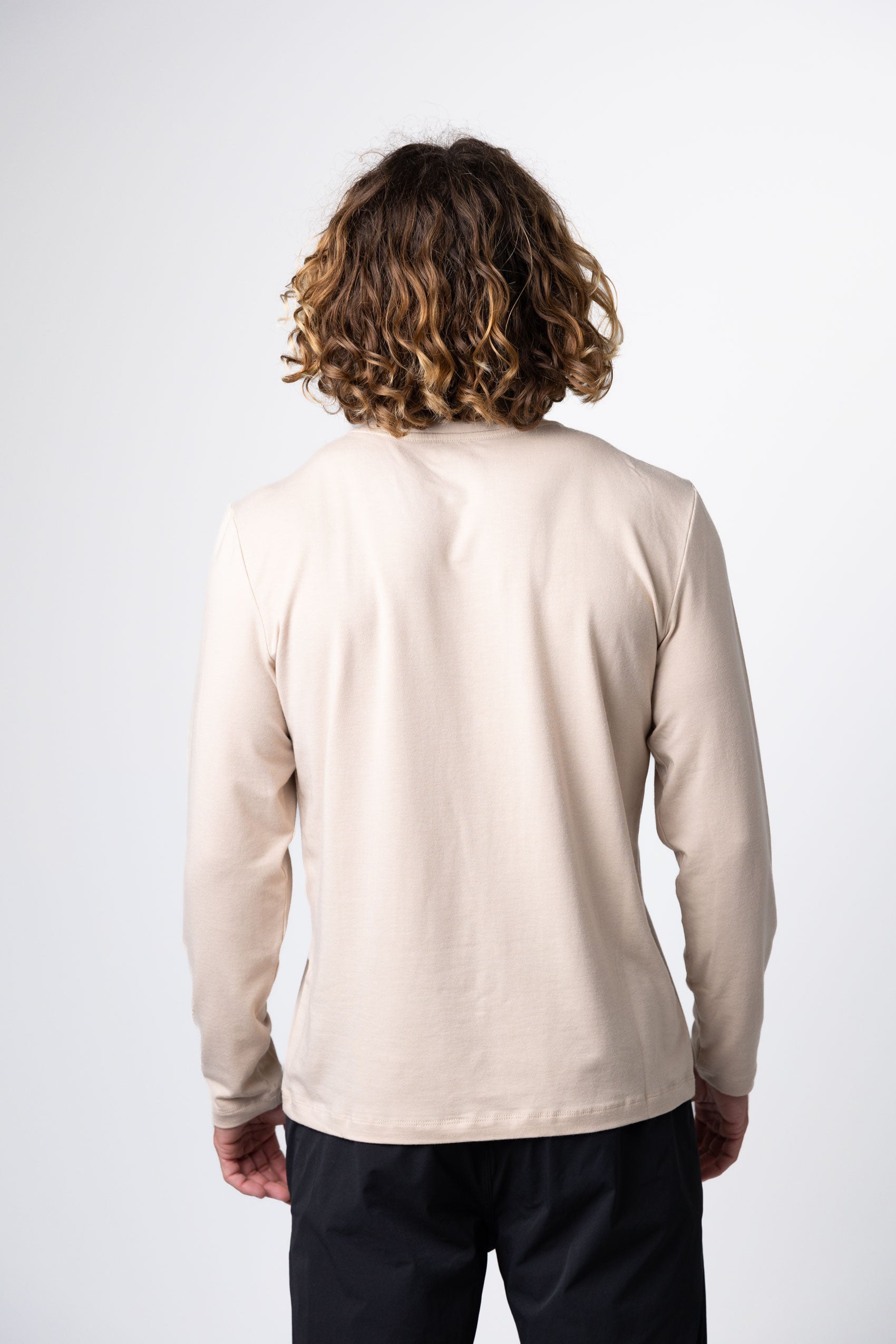 A person with shoulder-length curly hair stands facing away from the camera, wearing the Choona Versatile LS Henley with black pants against a plain white background.