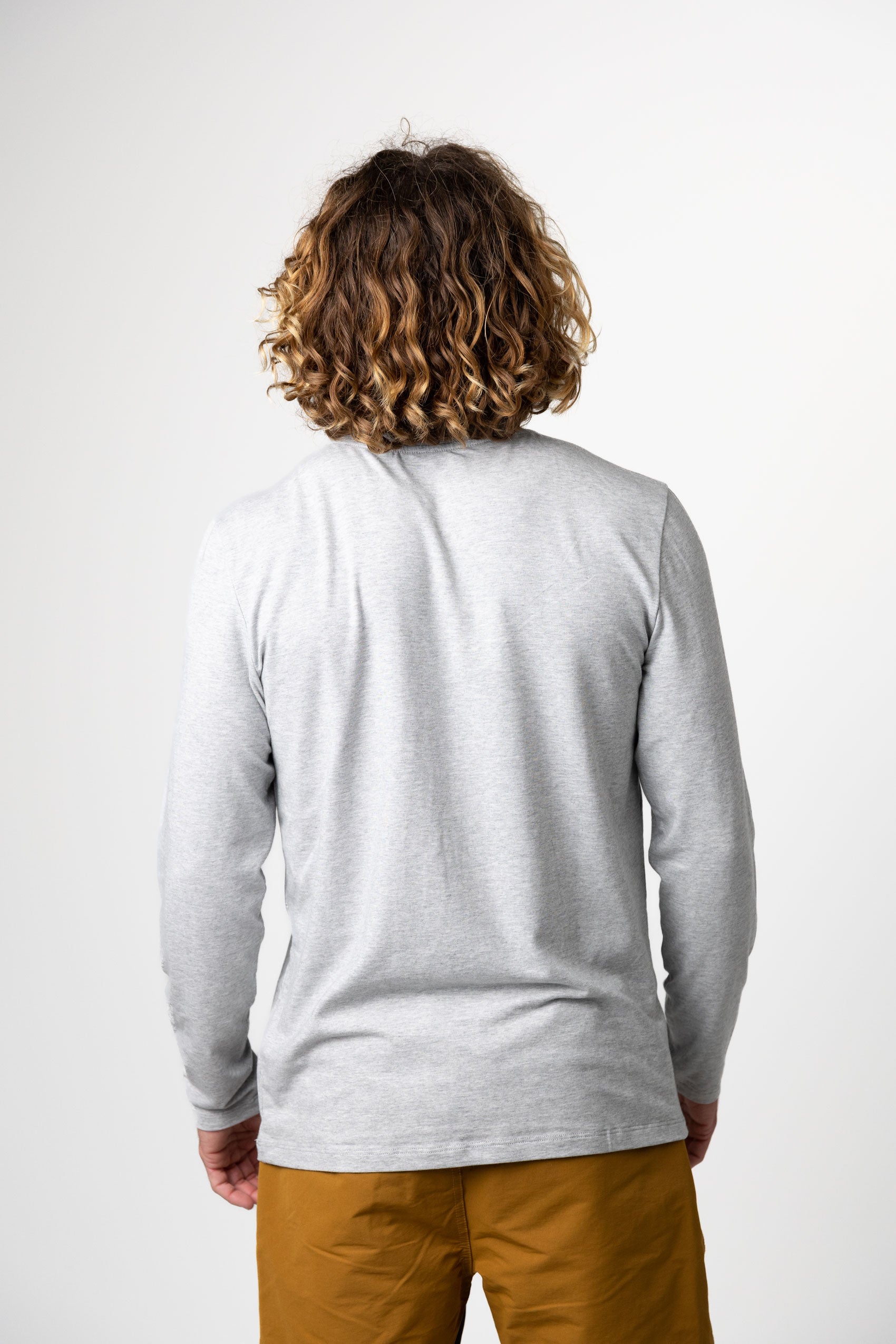 A person with curly, shoulder-length hair stands with their back to the camera, wearing the Choona Versatile LS Henley with mustard pants against a plain light background.