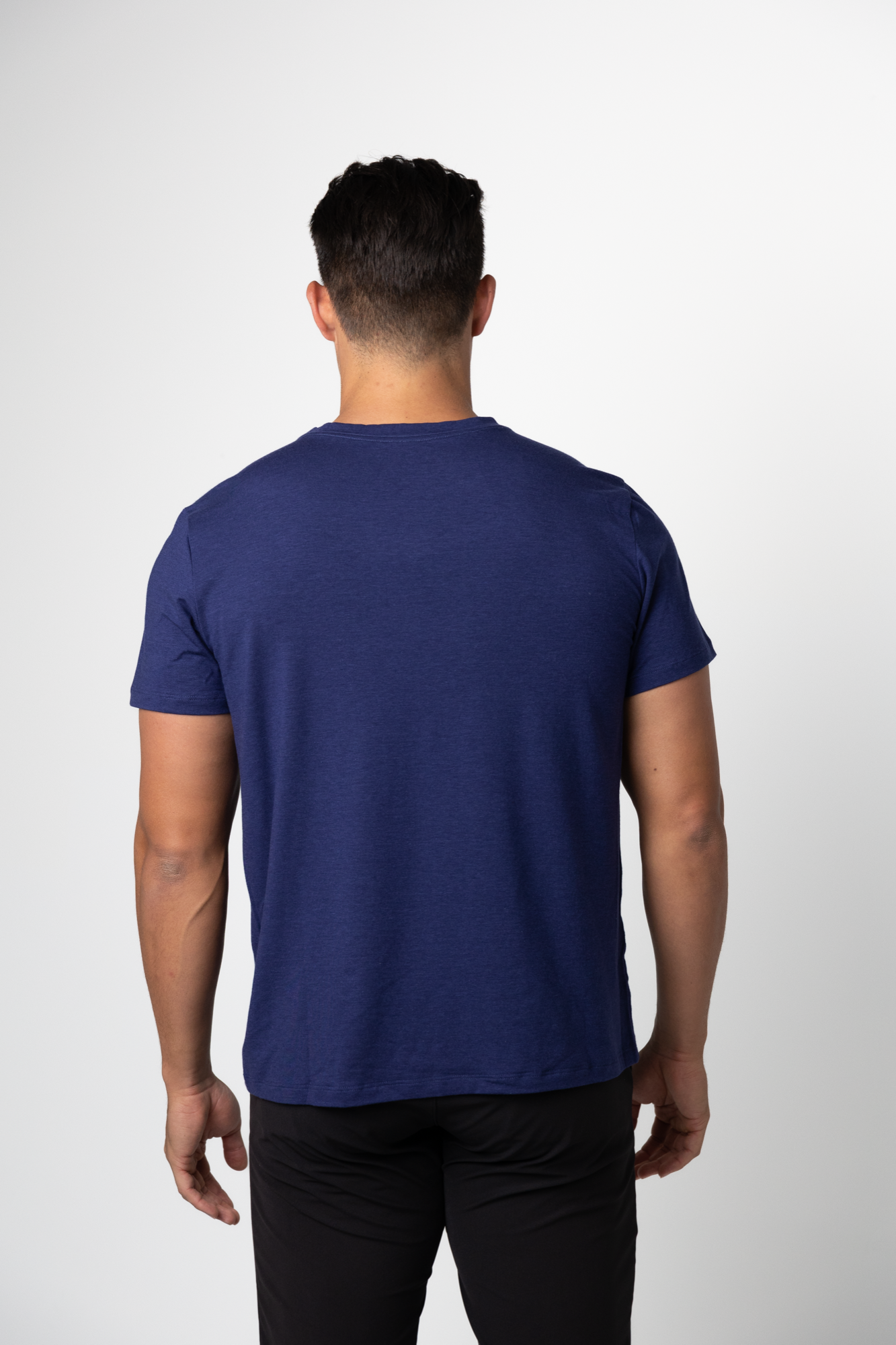 A man with short dark hair stands facing away in a blue Choona Panhandle Pocket Tee and black pants, set against a plain white background.