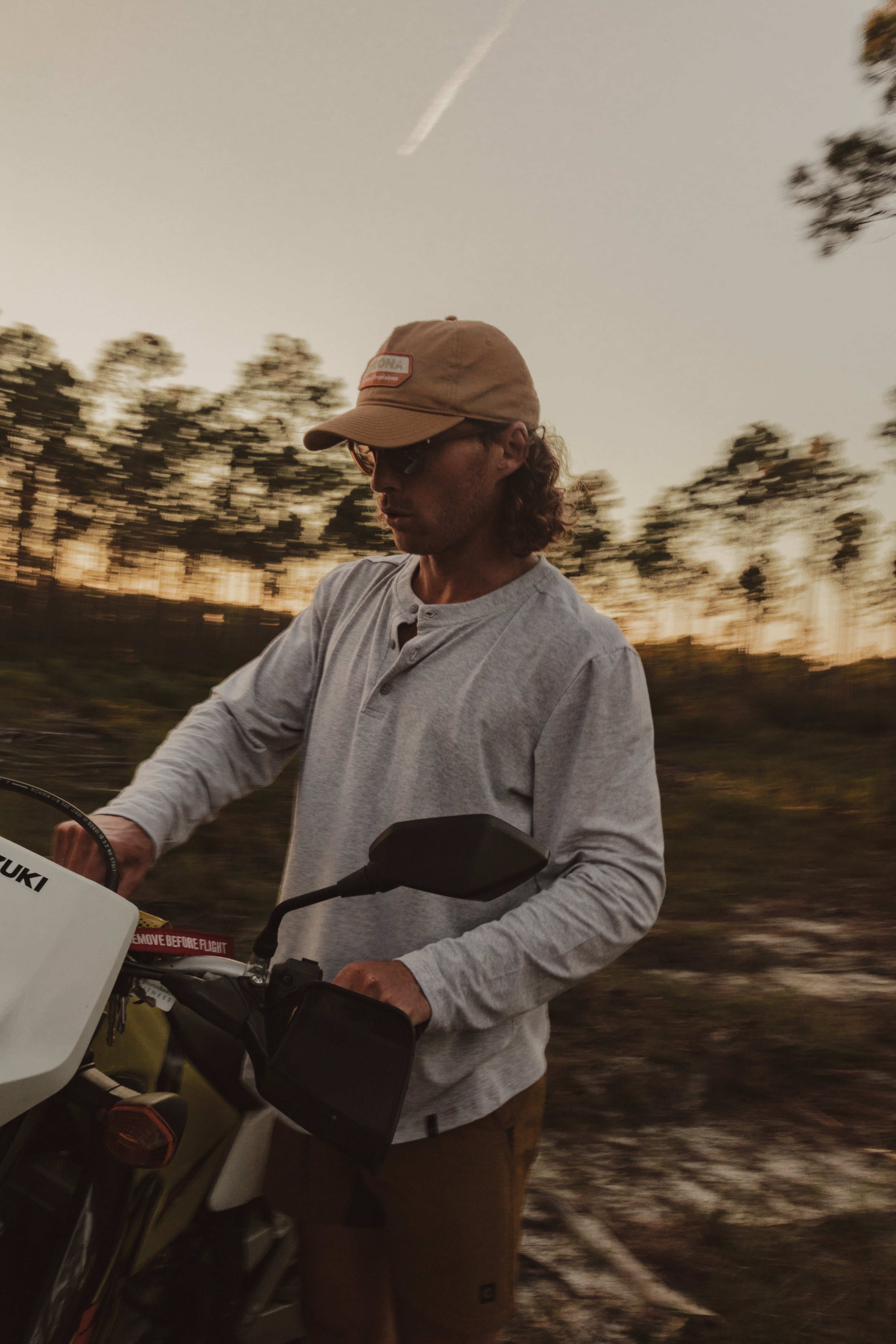 Wearing the Choona Versatile LS Henley, a person in a tan cap and sunglasses stands outdoors at sunset beside a white Suzuki motorcycle, embracing adventure among trees and clouds.