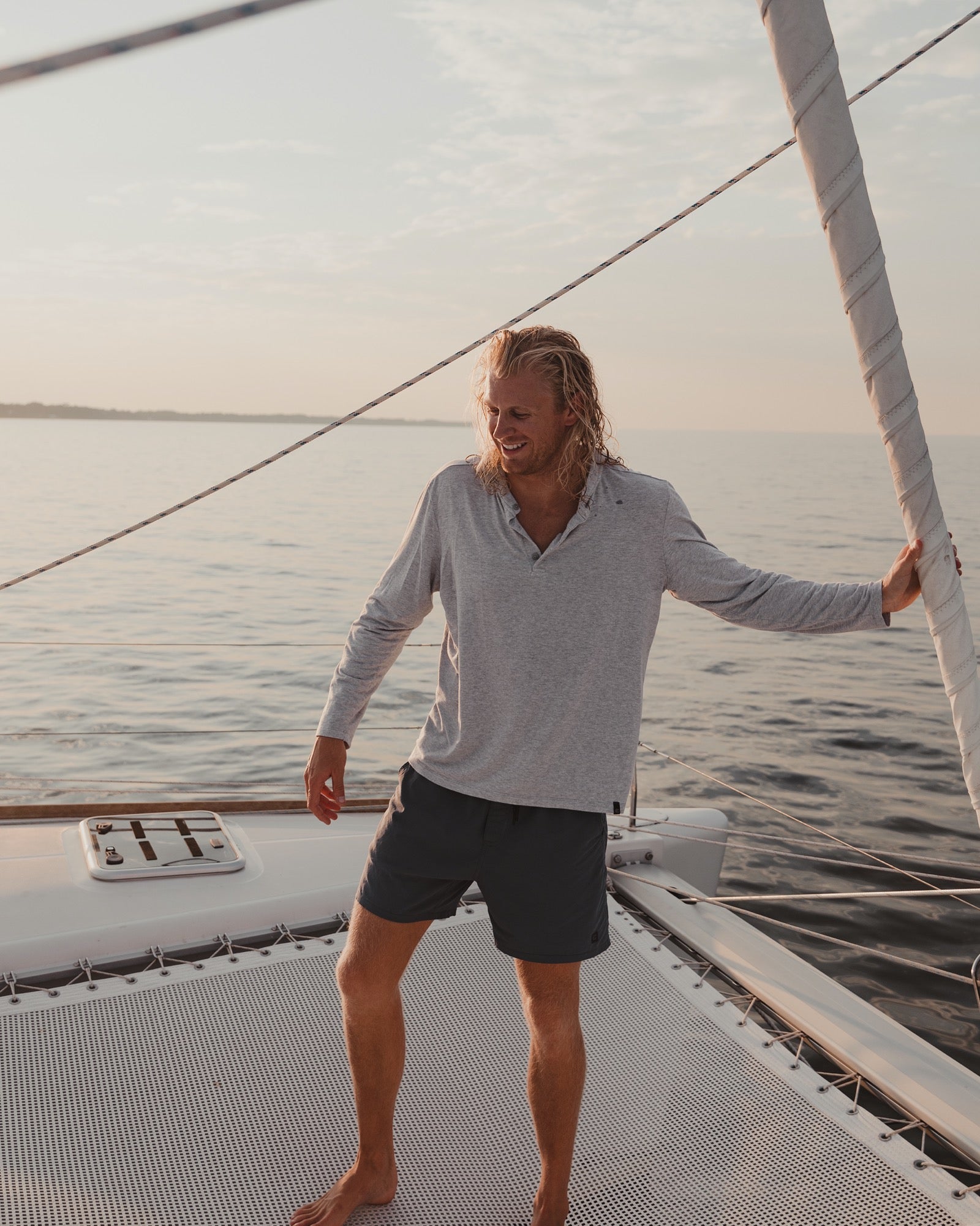 A man with long blond hair, barefoot on a sailboat, smiles while wearing the Choona Versatile LS Henley and dark shorts. He holds a rope as calm water and soft sunlight showcase his outdoor active lifestyle.