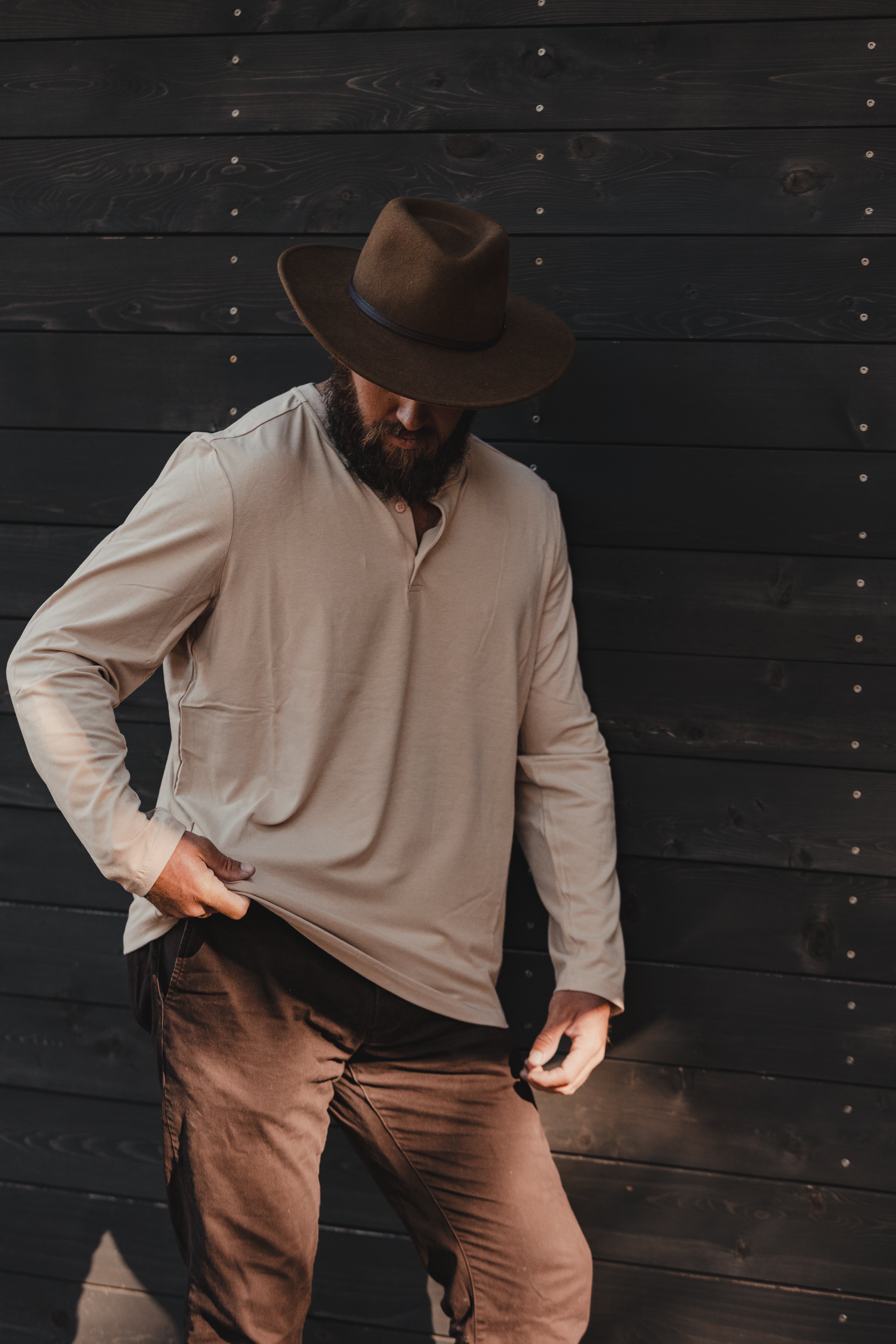 A bearded man wearing the Choona Versatile LS Henley, a wide-brimmed hat, and brown pants stands against a dark wooden wall—an effortless outleisure staple for an active outdoor lifestyle.