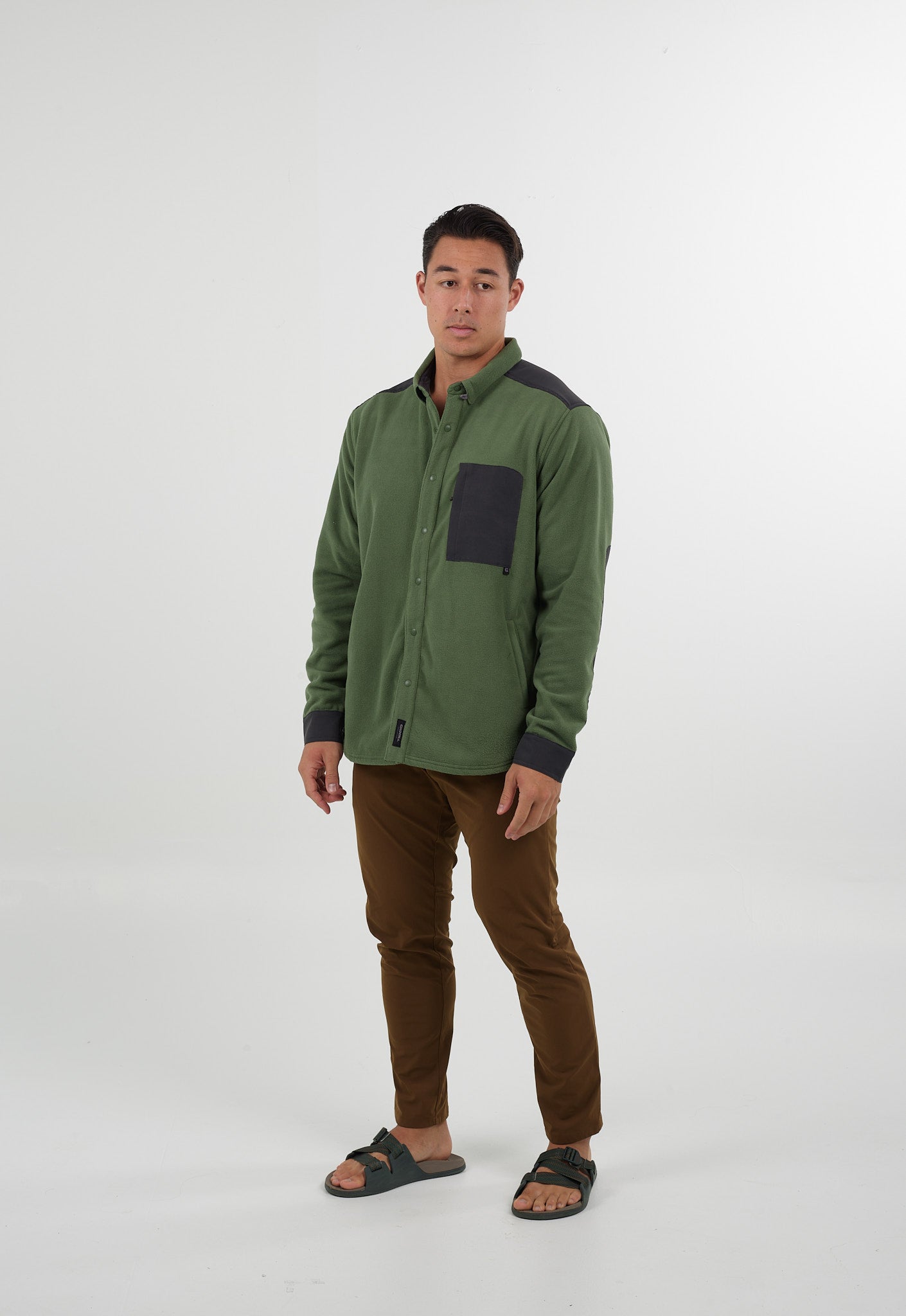 A man faces forward against a white background, wearing Choona’s green Skipper Shacket with a black chest pocket, plus brown pants and dark green sandals. Made from recycled polyester, this jacket is ideal for cold weather layering.
