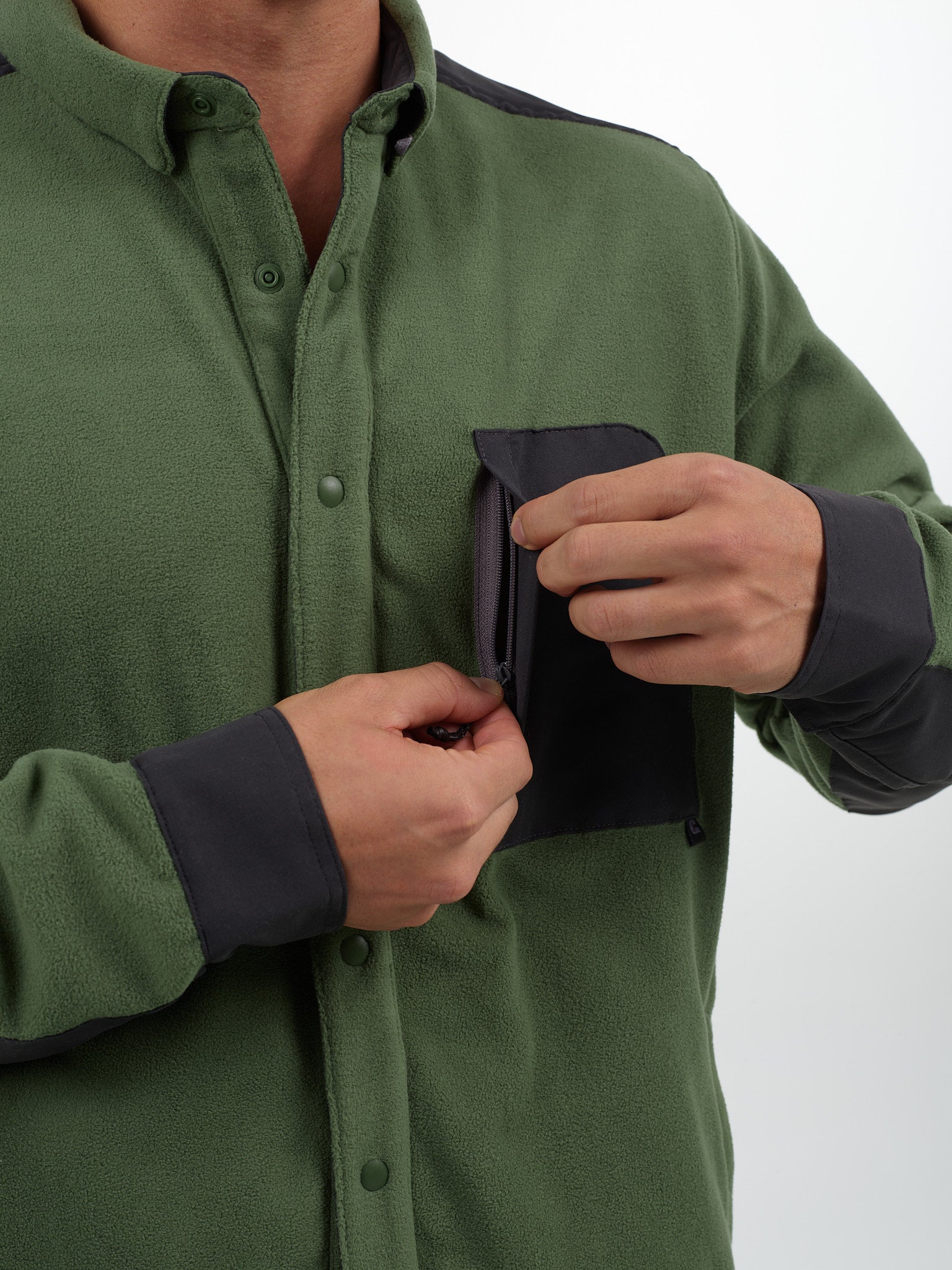 A person wearing Choonas green Skipper Shacket with dark gray accents is unzipping the small left chest pocket. Only the torso and hands are visible in this cold weather piece made from recycled polyester.